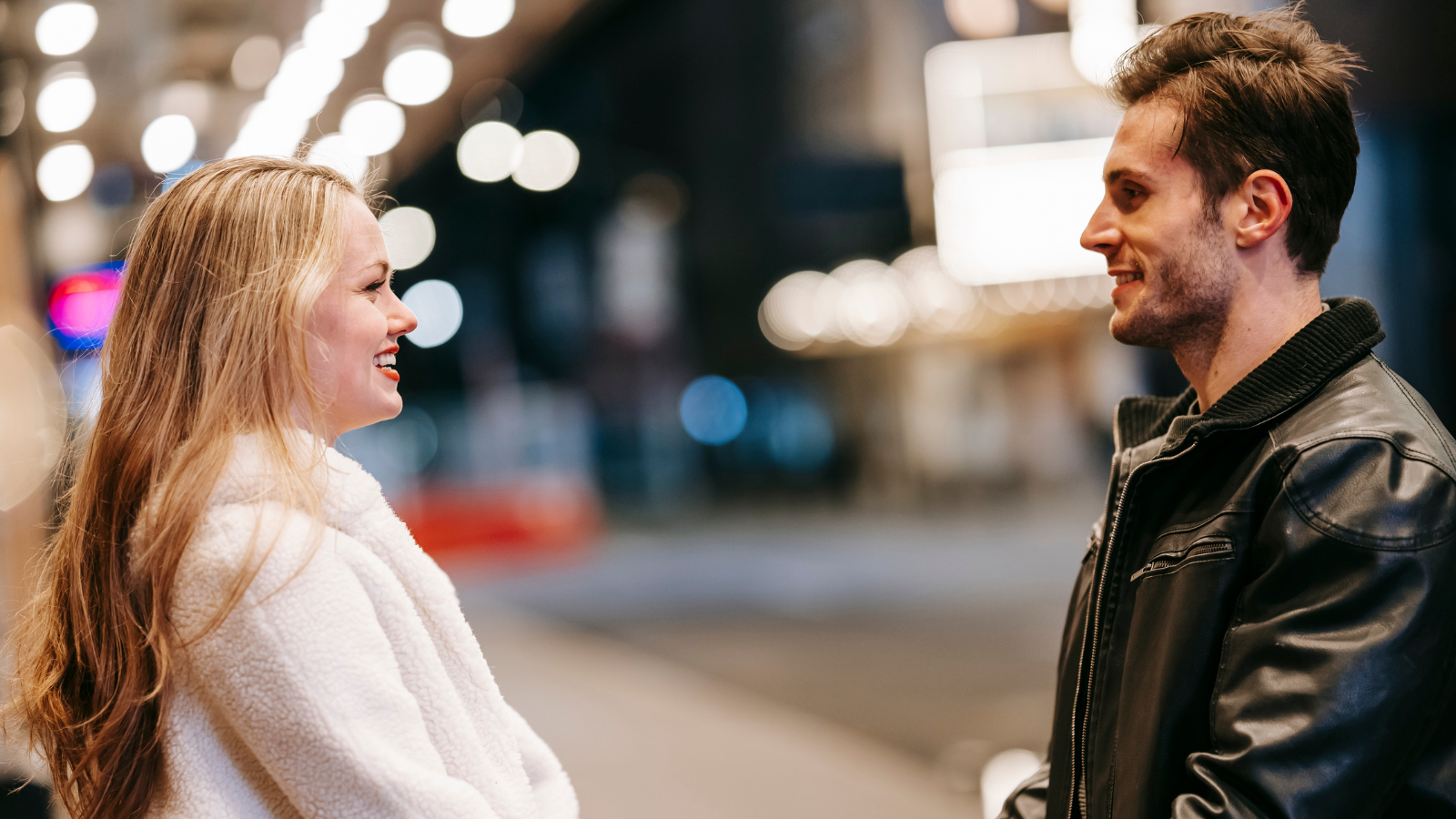 A man and woman smiling at each other on a nighttime street, exuding confidence and joy.