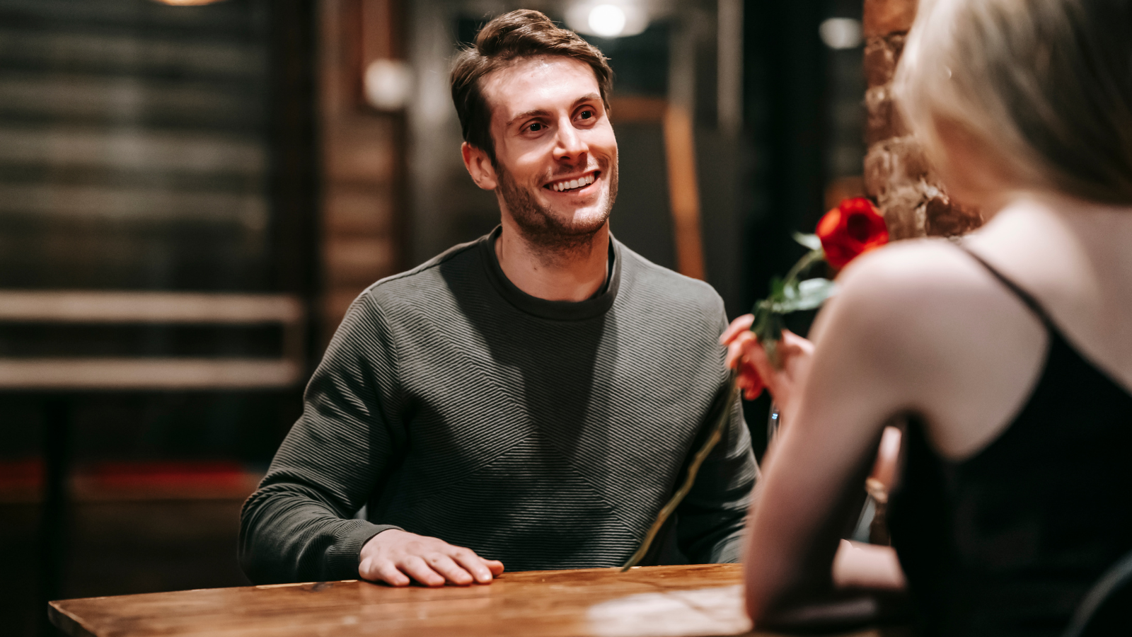 A man smiling and holding a rose, sitting at a table across from a woman in a dimly lit café.