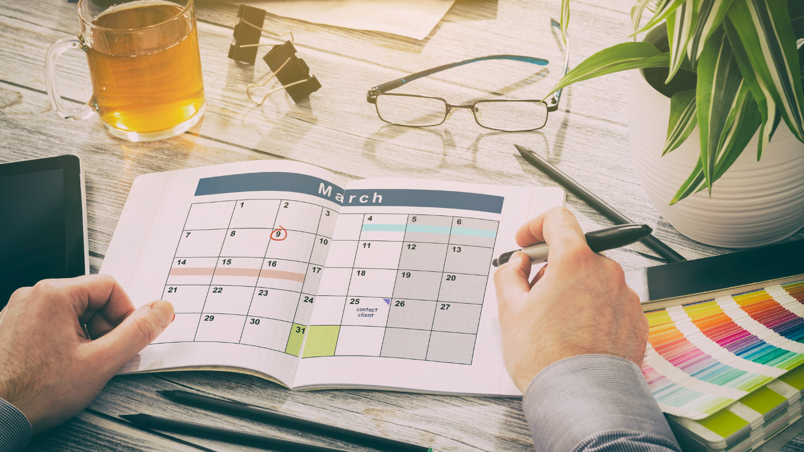 "A person organizing a schedule in a planner with a cup of tea and glasses on the table."