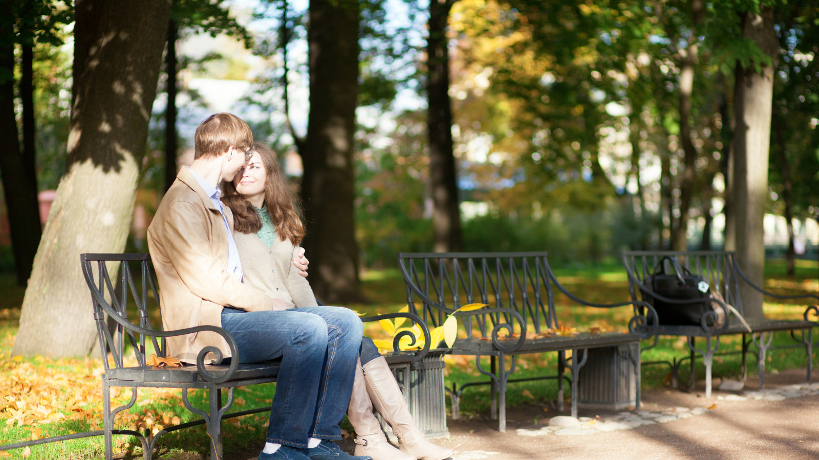 A couple sitting on a park bench, the man affectionately holding the woman as they smile at each other.