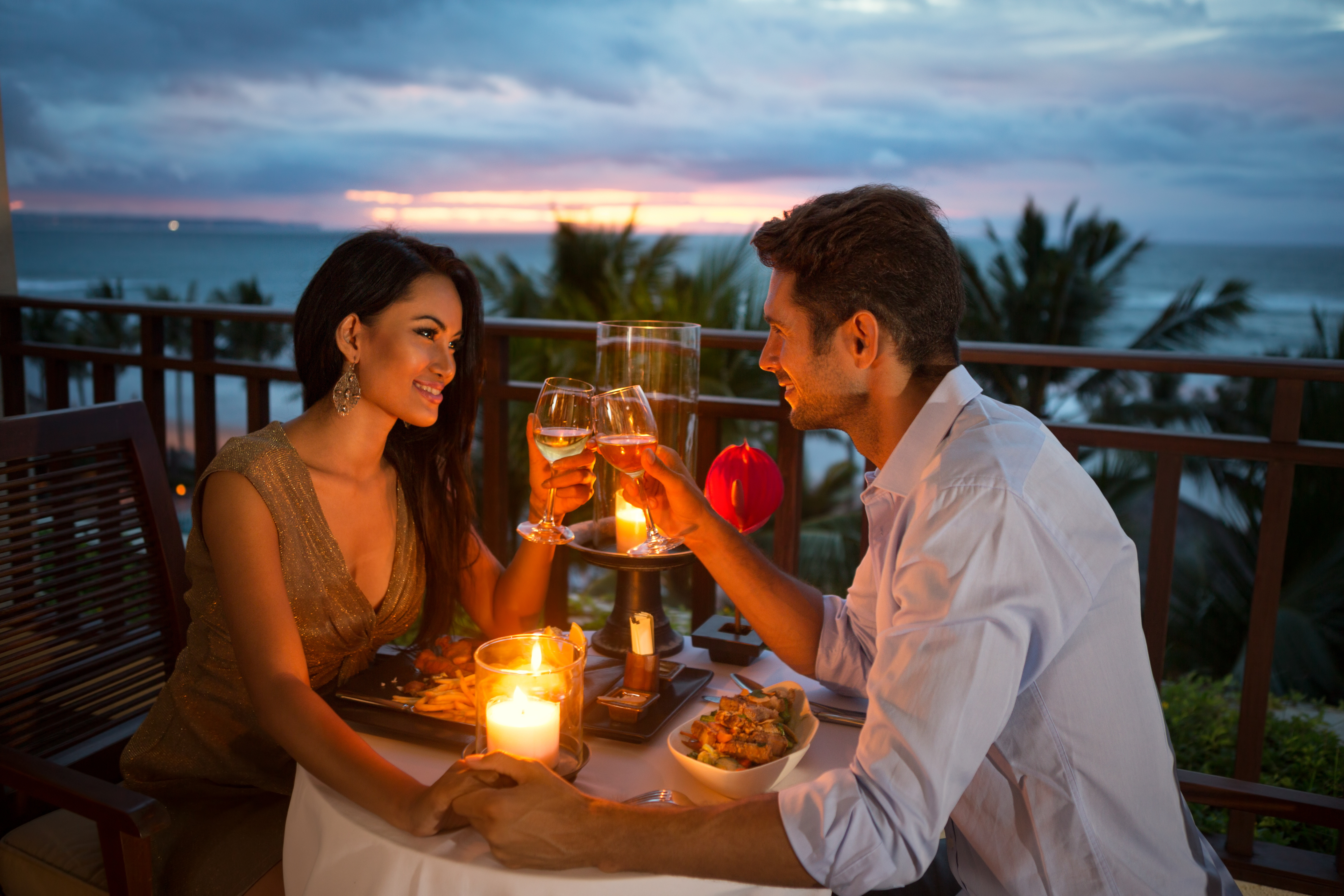 2 person enjoying a candlelit dinner on a seaside terrace with a romantic sunset in the background, smiling at each other and toasting.