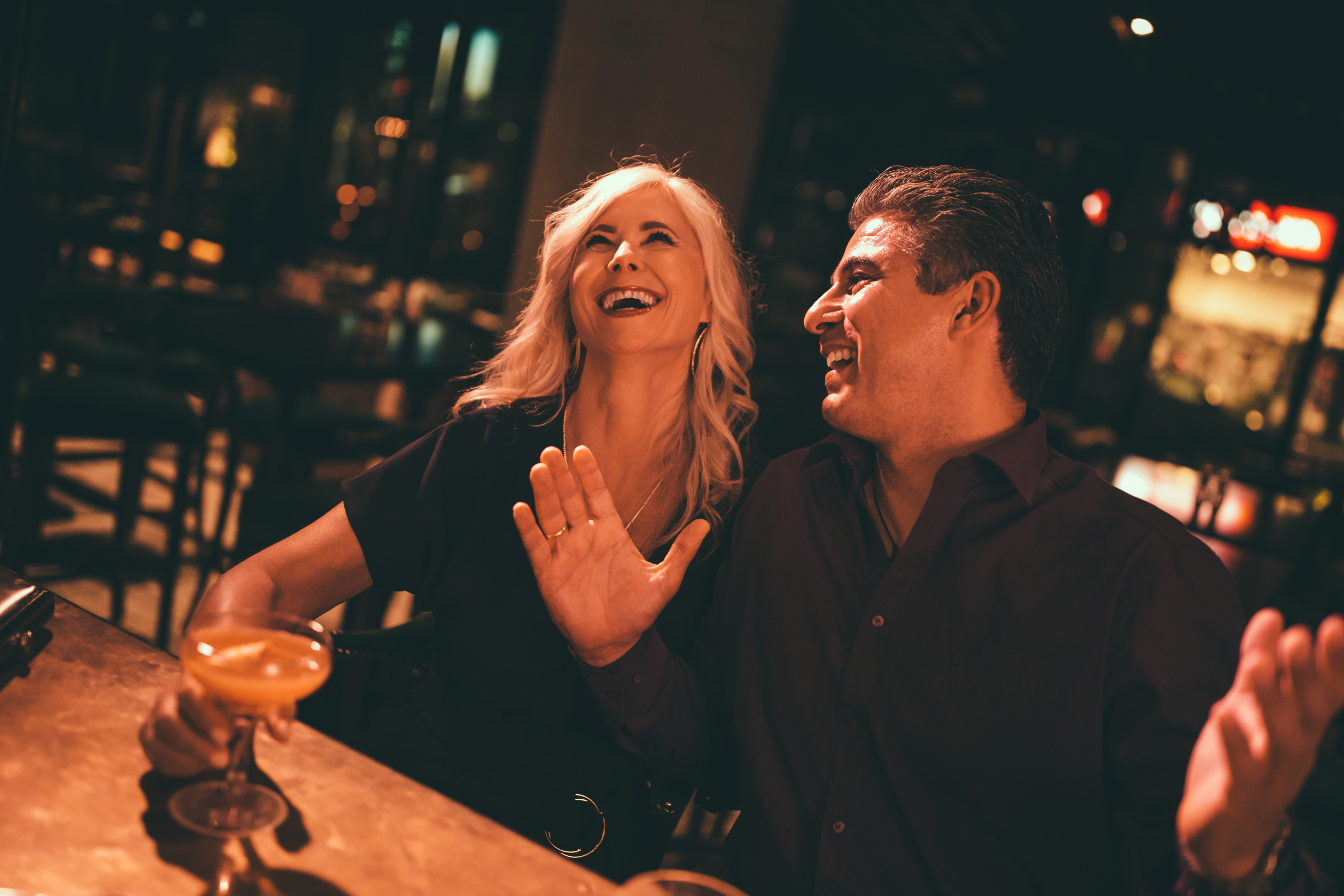 A middle-aged man and woman on a date at a bar, laughing and holding drinks. The warm lighting and their cheerful expressions highlight the moment, contrasting with the concept of dating fatigue.