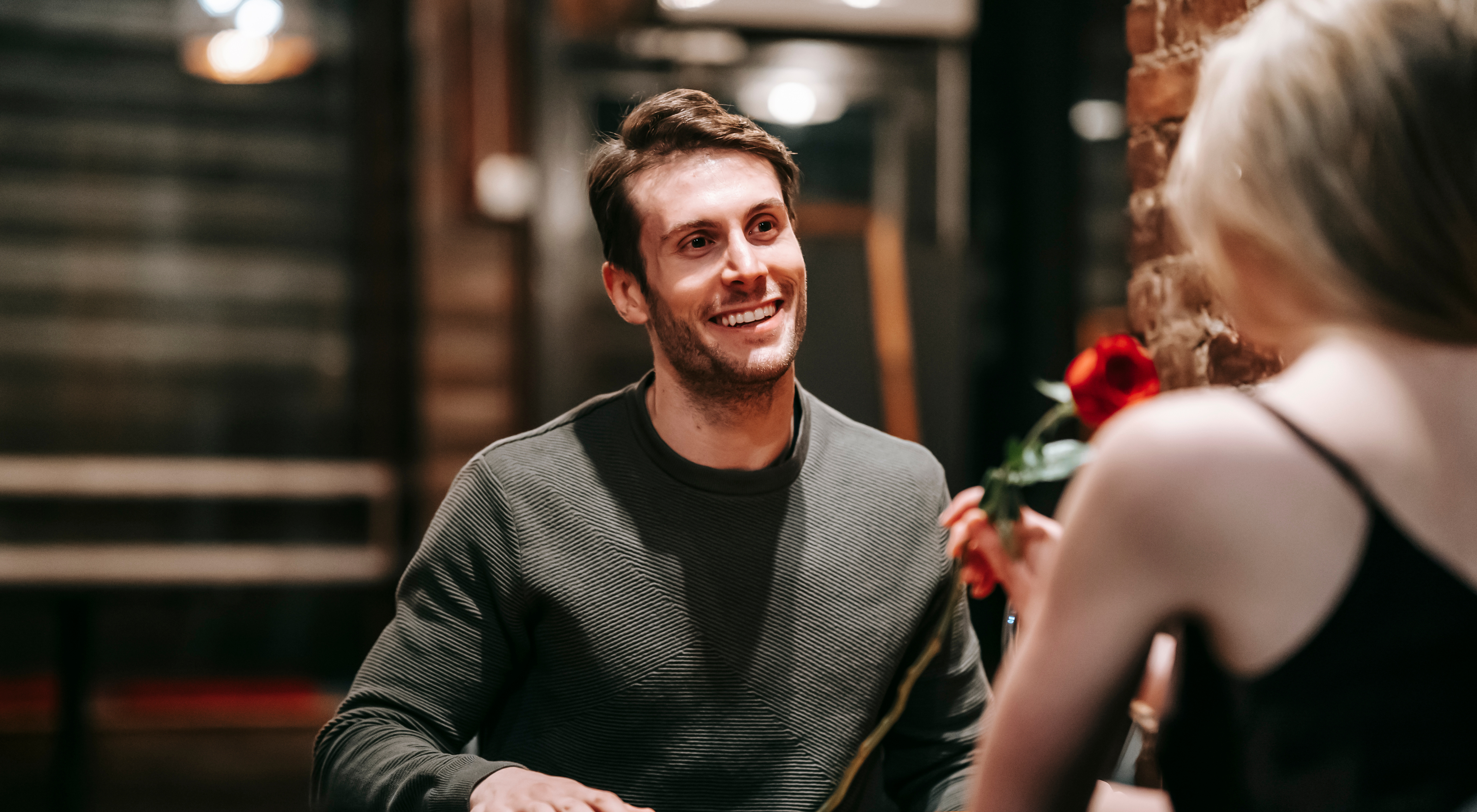 "A man smiling during a first date in a cozy restaurant, holding a red rose and engaging in light-hearted conversation."