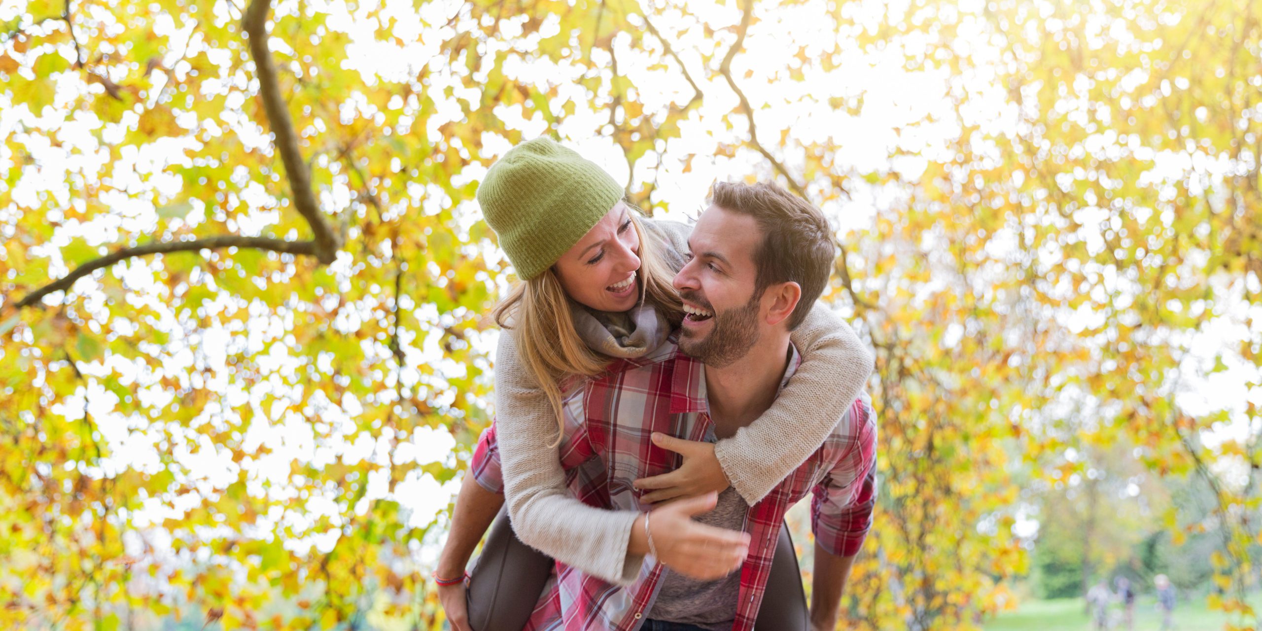 Happy couple laughing together in a park during autumn, with the woman playfully riding on the man's back