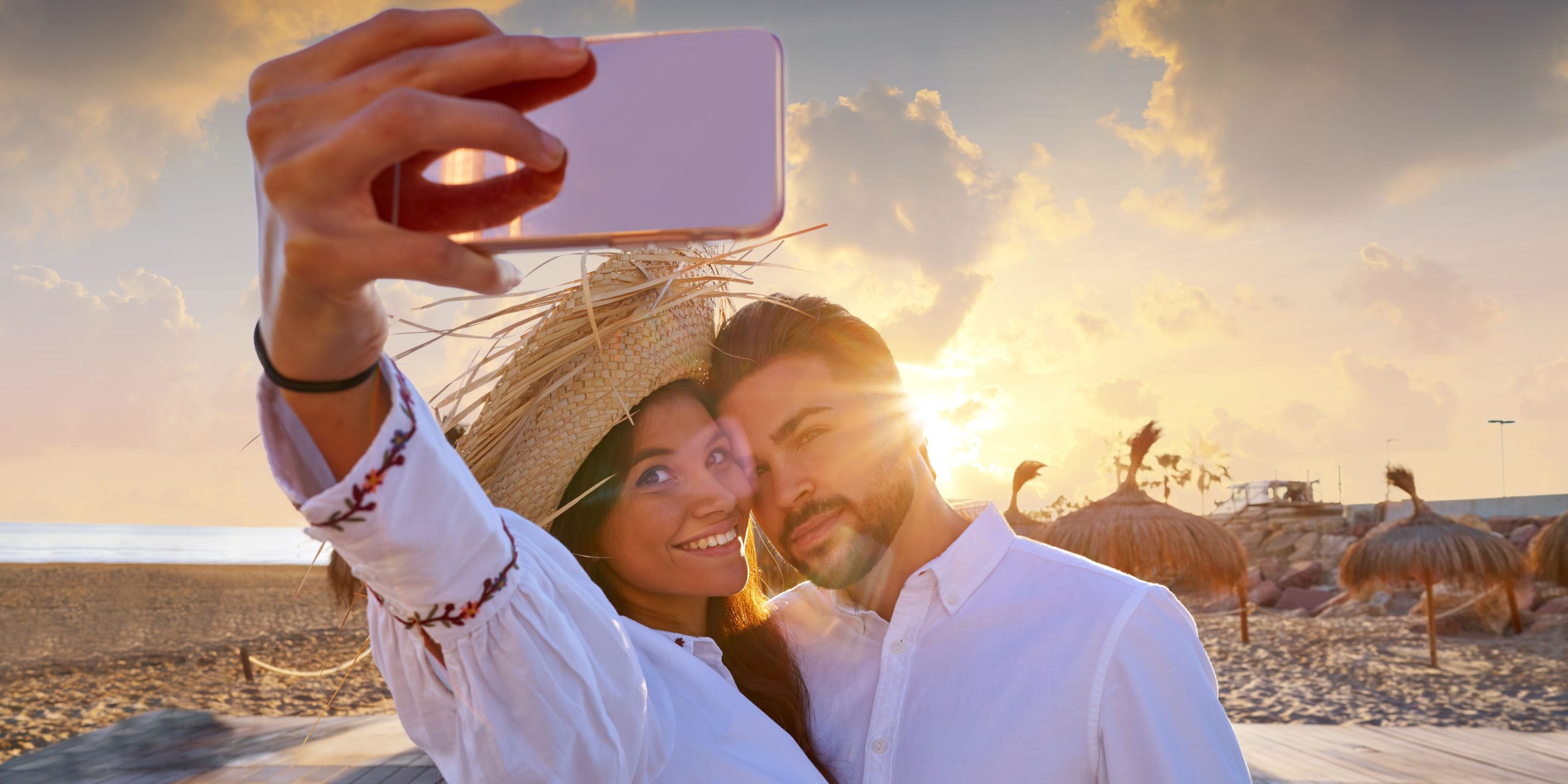 Smiling couple taking a selfie on a beach at sunset, enjoying a romantic moment with the sun setting behind them