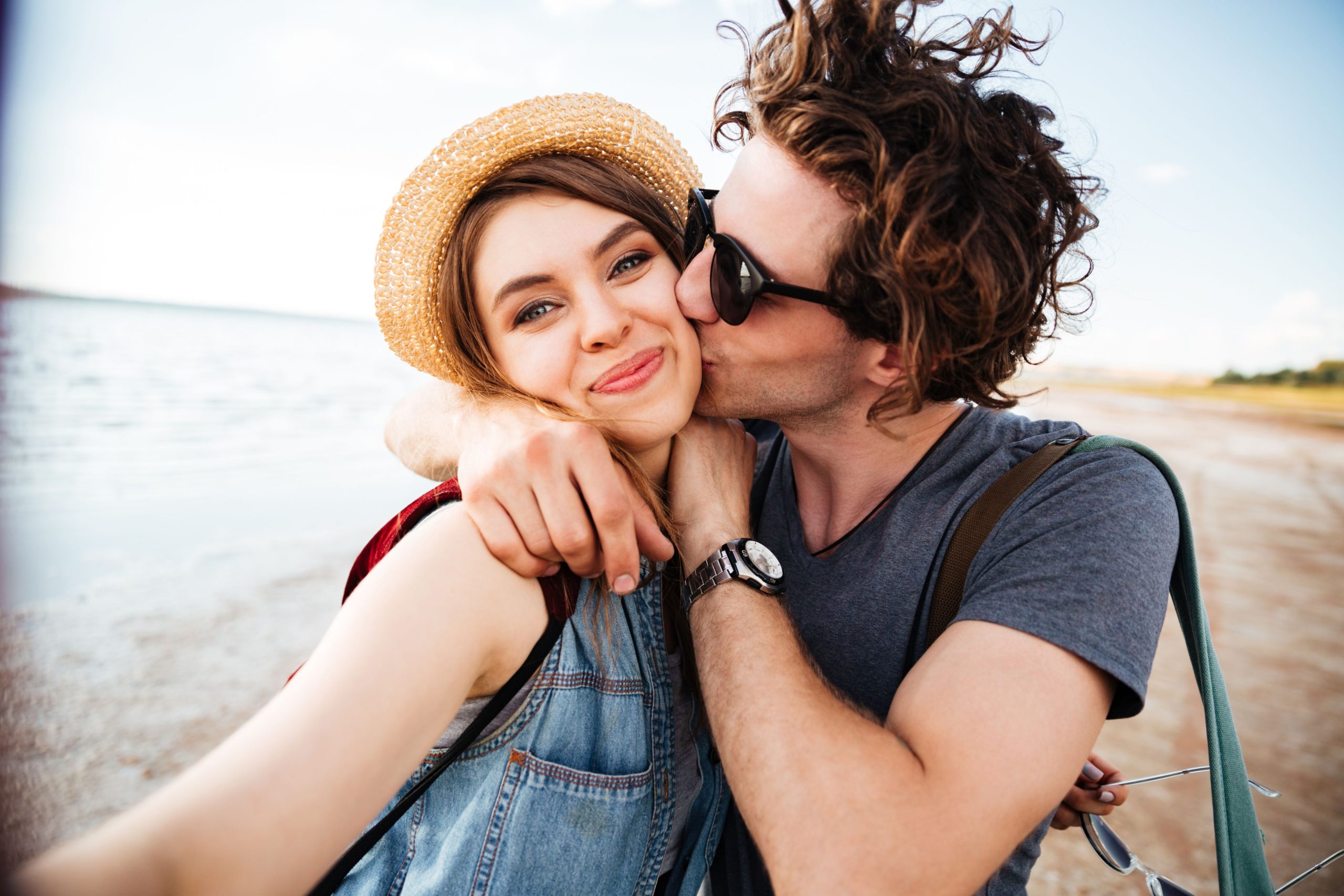 A couple at the beach, smiling and enjoying a sweet moment as the man kisses the woman on the cheek, capturing a carefree and romantic vibe.