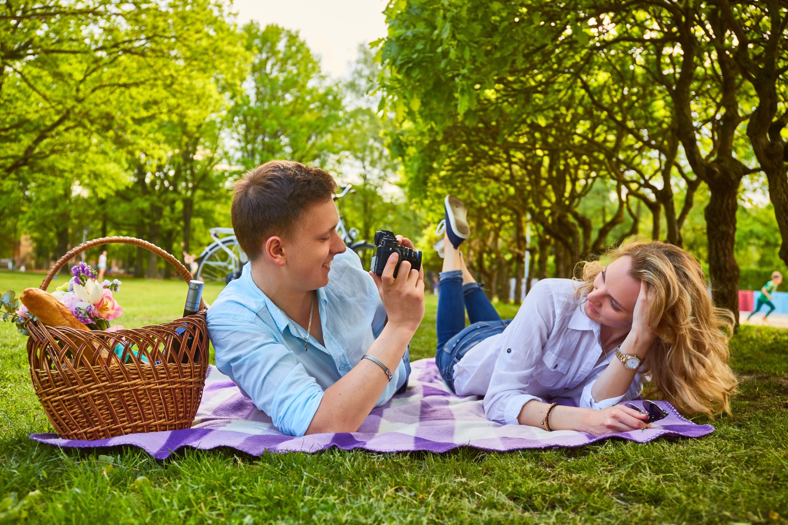 A couple enjoying a picnic on a sunny day in the park, lying on a blanket as the man shows the woman photos from a camera, illustrating a relaxed and joyful connection.