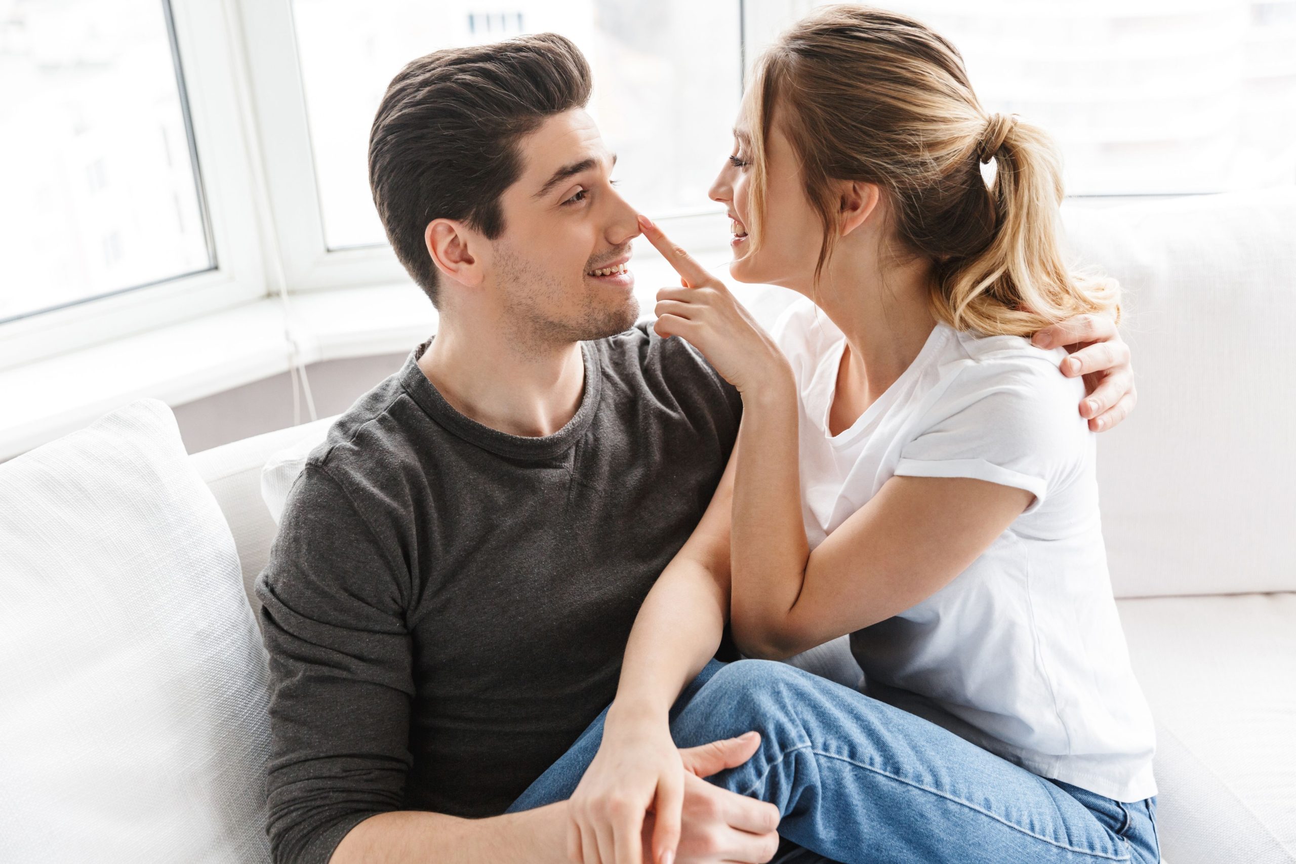 A couple sitting on a sofa at home, smiling as the woman playfully taps the man’s nose, showing a light-hearted, intimate moment.